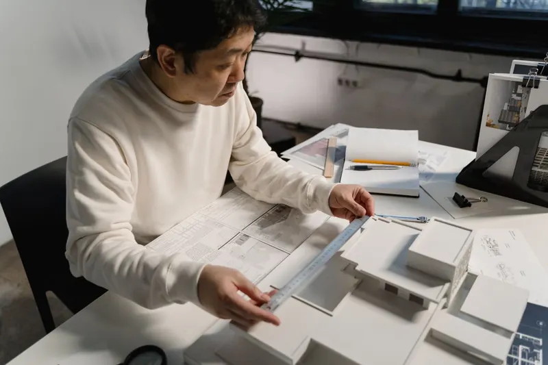 Arquiteto medindo uma maquete de casa em escala com uma régua sobre a mesa de projeto.