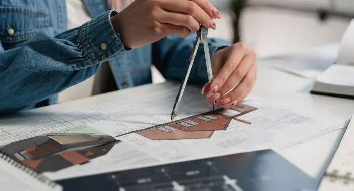 Mãos usando compasso sobre plantas e perspectiva de casa em mesa de projeto