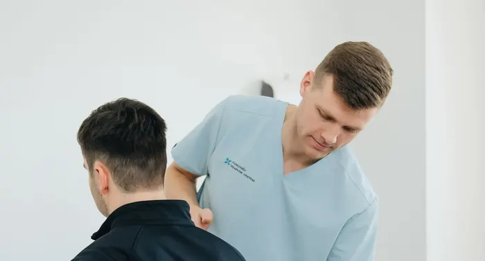 Fisioterapeuta de uniforme azul-claro avaliando as costas de um paciente em sala de atendimento
