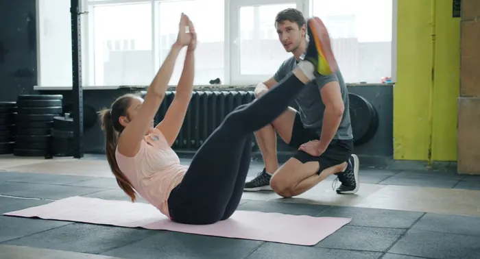 Treinador ajoelhado orienta mulher fazendo abdominal sobre colchonete em sala de treino