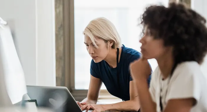 Duas colegas colaborando em notebooks em uma mesa junto à janela de um escritório