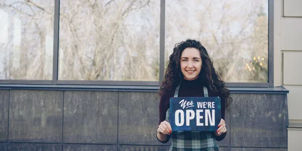 Empreendedora sorrindo enquanto segura uma placa de aberto na frente da loja