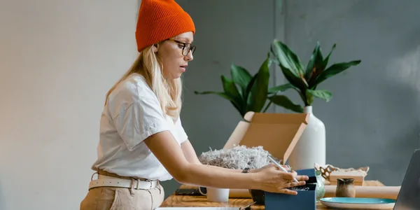 Mulher de gorro laranja embalando encomendas em uma mesa de madeira