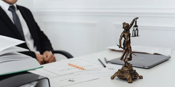 Estatueta da justiça sobre a mesa do escritório de advocacia, com documentos e um advogado sentado ao fundo.