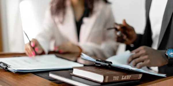 Duas pessoas sentadas à mesa de um escritório de advocacia, com um livro de leis e documentos entre elas.