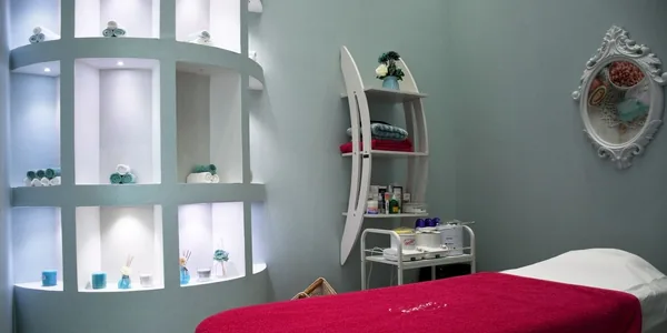 Sala de atendimento de clínica de estética com maca preparada, carrinho auxiliar e prateleiras iluminadas com os produtos