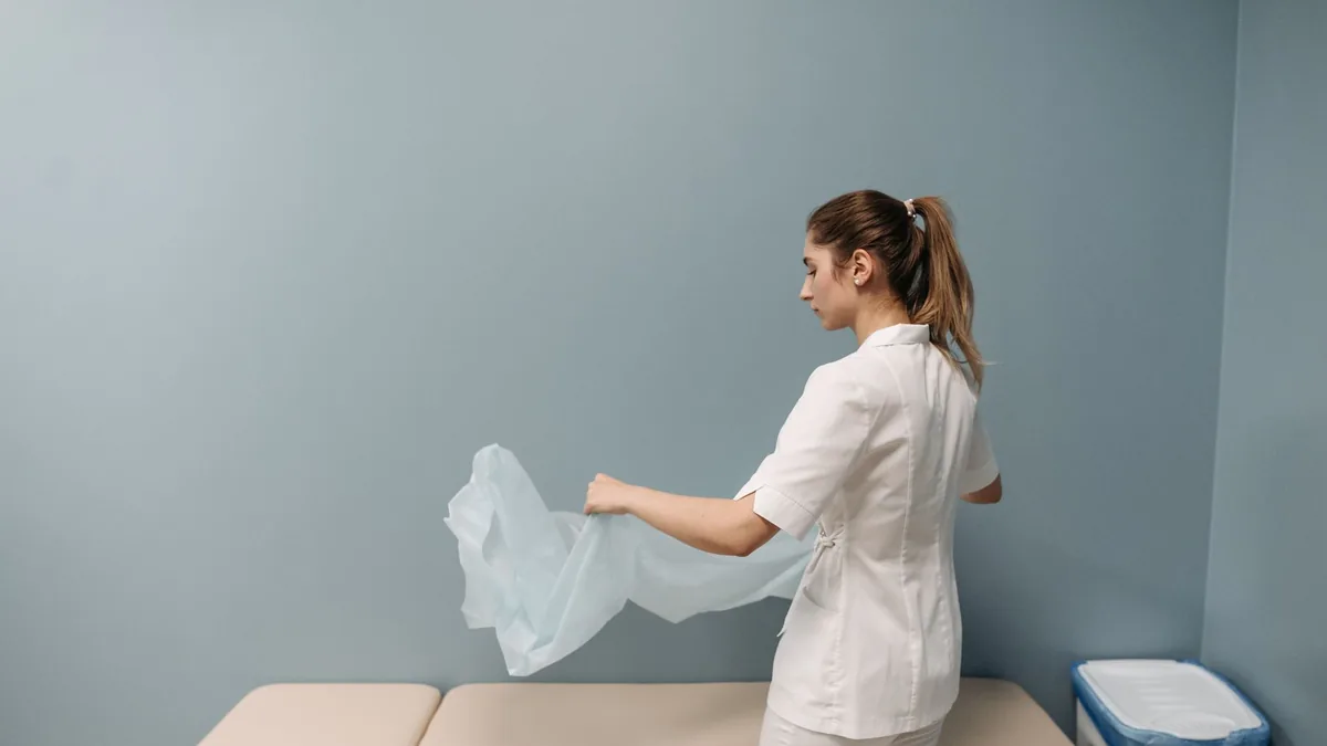 Profissional de uniforme branco estendendo o lençol sobre a maca antes do próximo atendimento