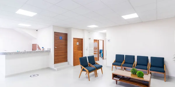 Sala de espera clara de uma clínica médica, com o balcão da recepção ao fundo e uma fileira de cadeiras azuis