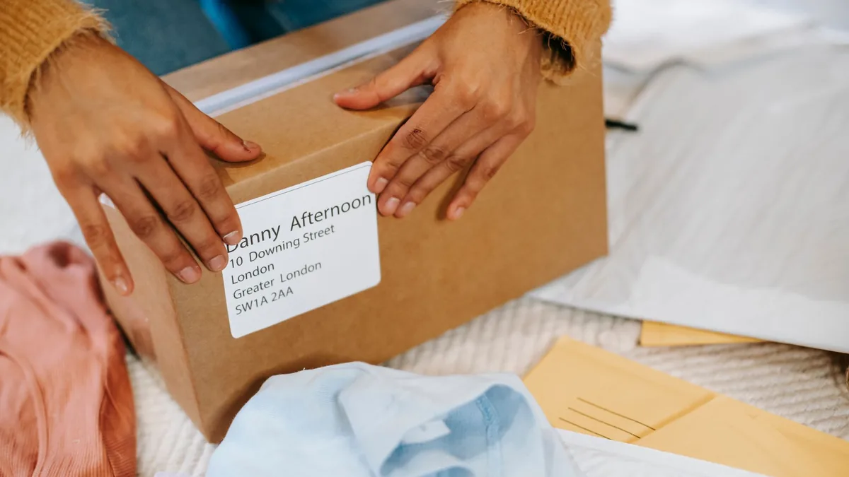 Mãos fechando uma caixa de papelão com etiqueta de endereço, com as peças e os envelopes sobre a mesa