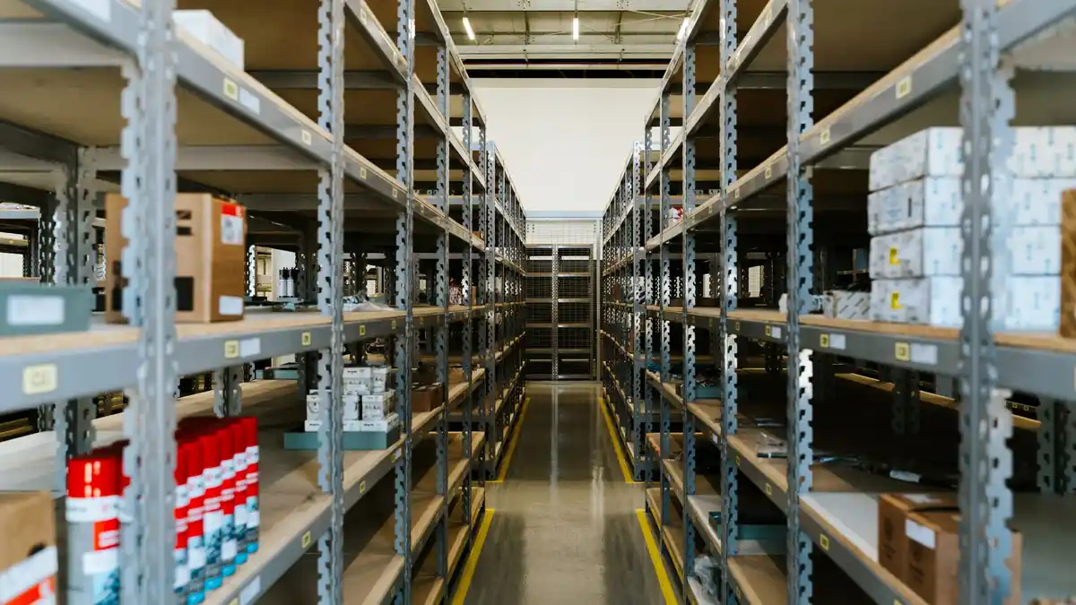 Corredor de um estoque com prateleiras metálicas de ambos os lados, cheias de caixas e produtos etiquetados