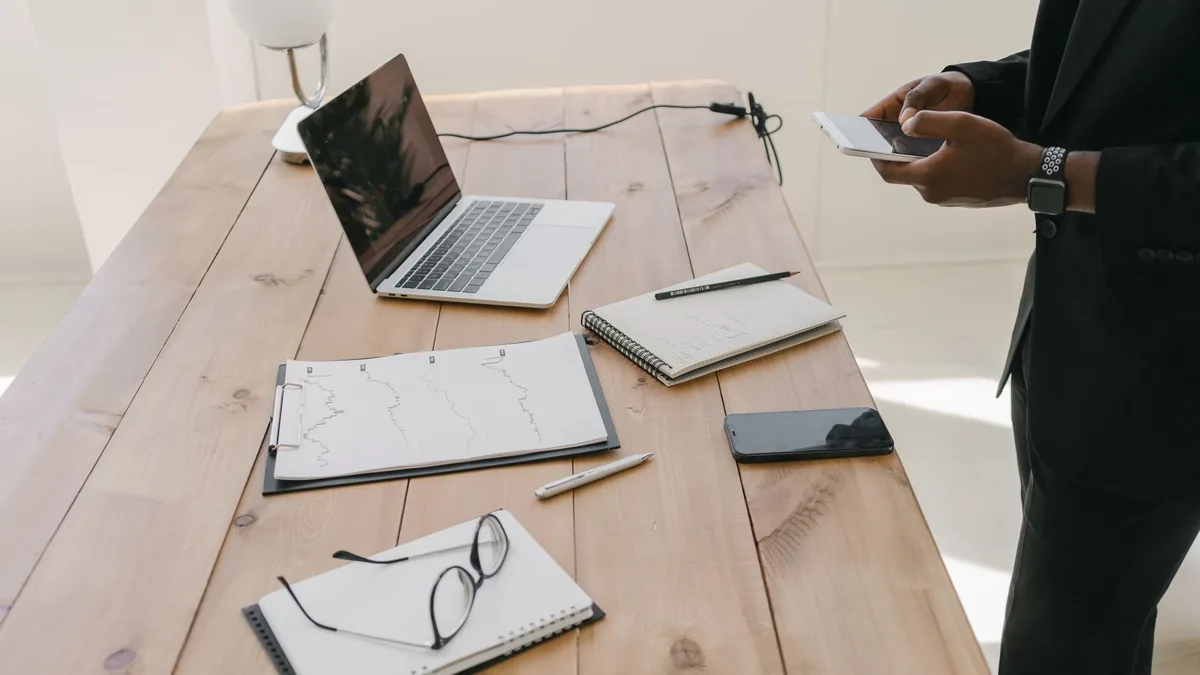 Mesa de madeira com notebook aberto, cadernos de anotações e uma pessoa com o celular na mão