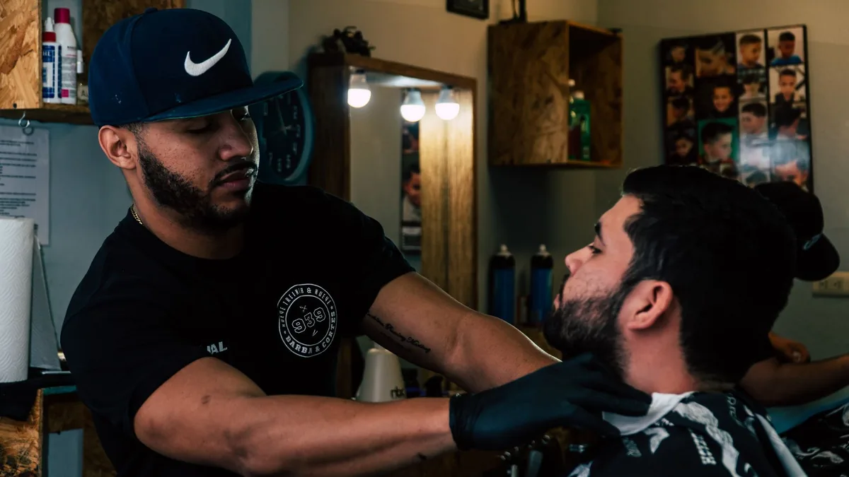 Barbeiro de boné reclinando a cadeira para atender um cliente, com os espelhos e as fotos da barbearia ao fundo