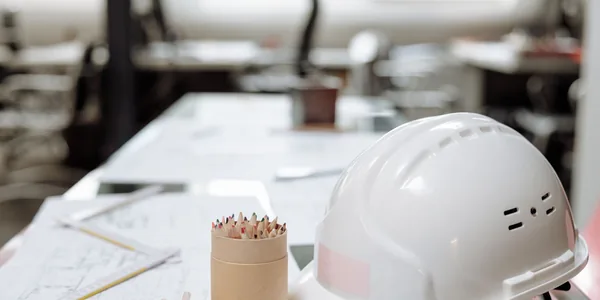 Capacete branco de obra sobre a mesa de projeto, ao lado de plantas impressas e de um porta-lápis.