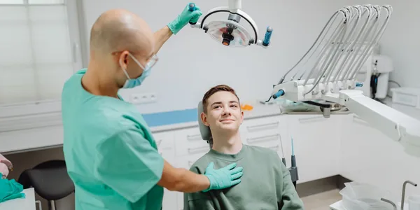Dentista de jaleco verde ajustando o refletor sobre um paciente sentado na cadeira do consult&oacute;rio.