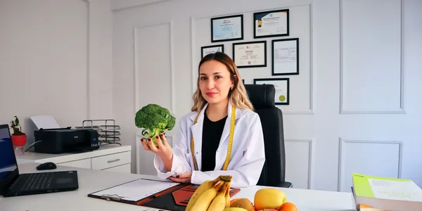 Nutricionista de jaleco branco sentada à mesa do consultório, com frutas e legumes à frente e diplomas na parede.