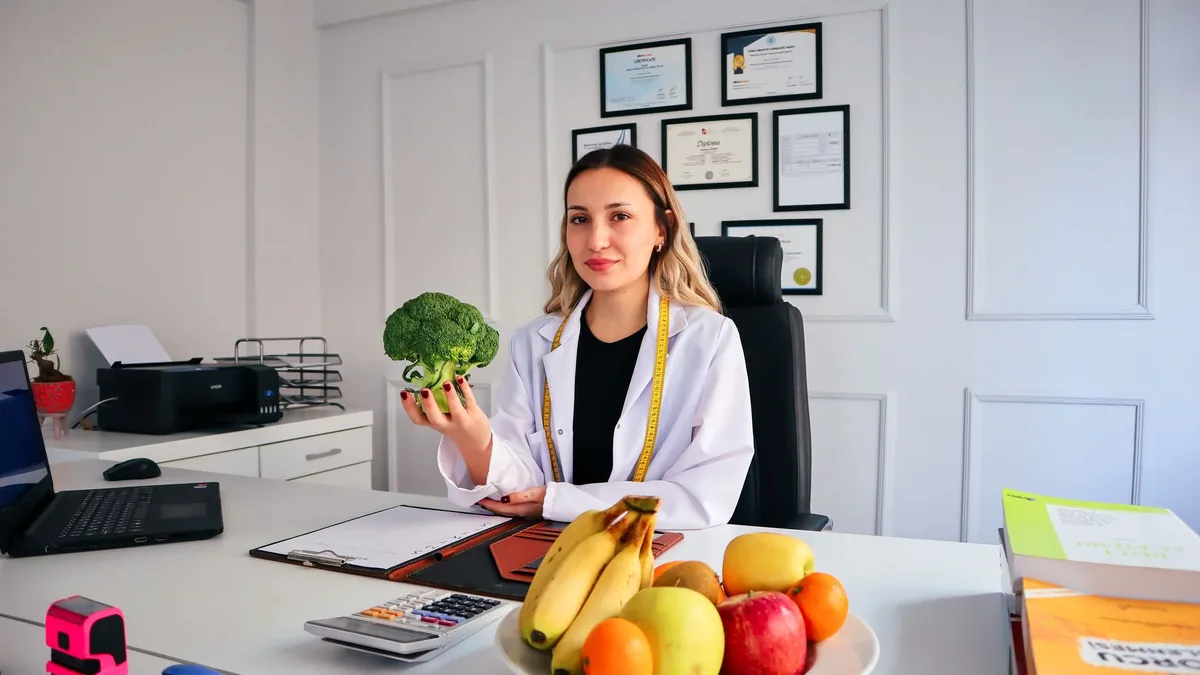 Nutricionista no consultório segurando um brócolis ao lado de uma fruteira sobre a mesa.