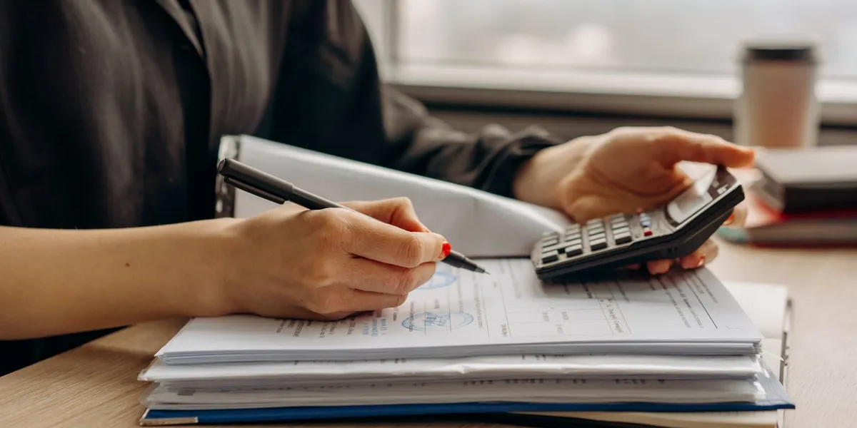 Pessoa calculando com uma calculadora sobre pastas e documentos financeiros em uma mesa de escritório.