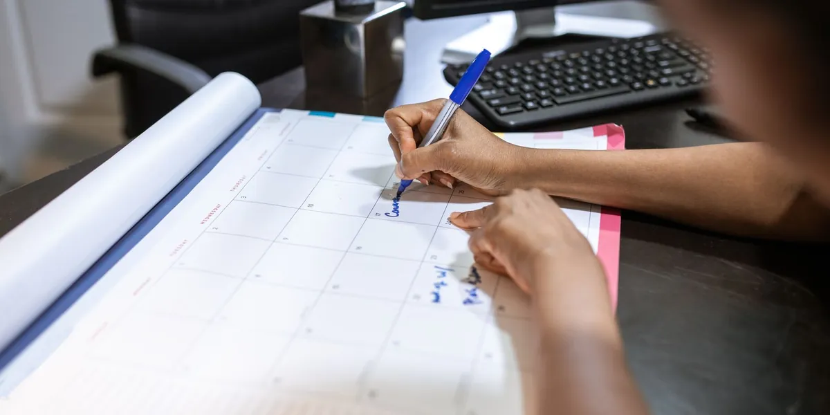 Mãos de uma pessoa escrevendo com caneta em um calendário de mesa, em um ambiente de escritório.