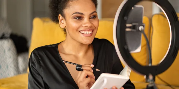 Criadora de conteúdo sorrindo enquanto grava um vídeo com o celular no tripé e o aro de luz ligado.