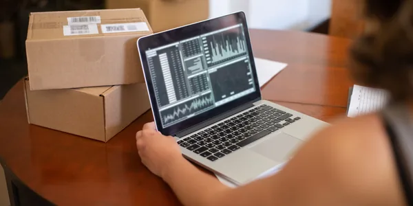 Caixas de papelão etiquetadas ao lado de um notebook com gráficos de vendas, na mesa de quem prepara os pedidos.