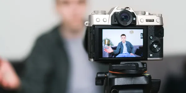 Câmera fotográfica sobre tripé, com a tela virada mostrando um criador gravando um vídeo ao fundo