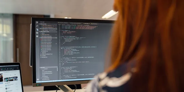Desenvolvedora de costas em frente a um monitor com código aberto e um notebook ao lado