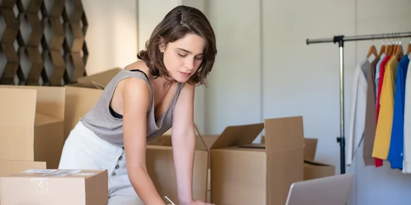 Lojista anotando pedidos sobre a mesa, cercada de caixas de papelão, com notebook e arara de roupas ao fundo