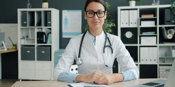 M&eacute;dica de jaleco branco sentada &agrave; mesa do consult&oacute;rio, com prancheta de documentos e notebook