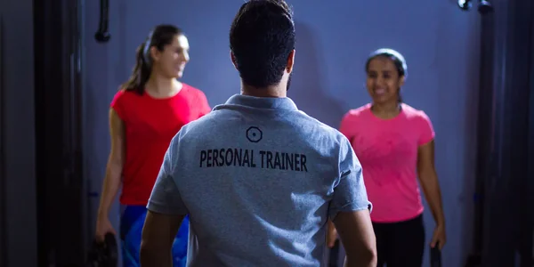 Personal trainer de camisa cinza escrita Personal Trainer orientando duas alunas na academia