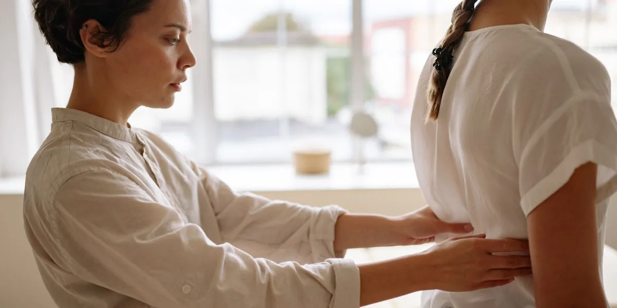 Fisioterapeuta com as mãos na lombar de uma paciente em pé, avaliando a postura em sala clara.