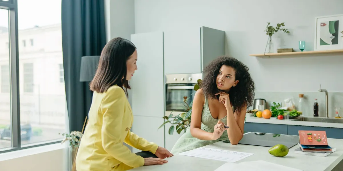 Nutricionista conversando com uma cliente apoiada na bancada da cozinha, com frutas e um caderno sobre o balcão.