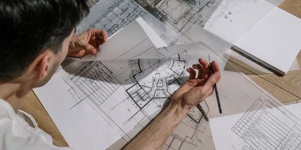Arquiteto desenhando com lápis sobre plantas técnicas espalhadas na mesa de trabalho.