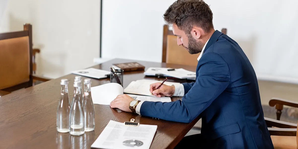 Homem de terno escrevendo em um caderno sobre a mesa de madeira de uma sala de reunião.