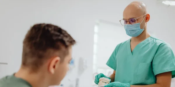 Dentista de jaleco verde e máscara mostrando um modelo de arcada dentária a um paciente no consultório