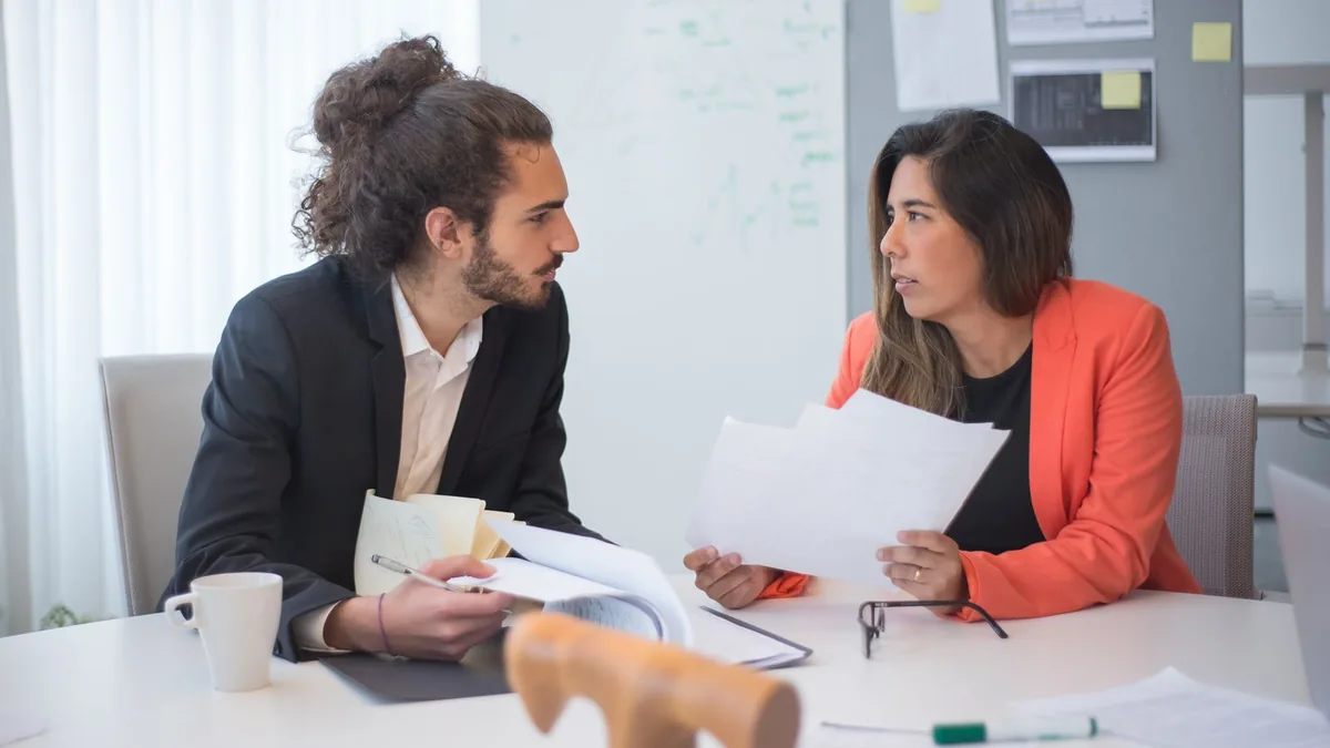 Duas pessoas conversando sobre documentos em uma mesa de escritório com café ao lado