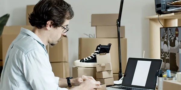 Lojista embalando pedidos em caixas de papelão ao lado do notebook no estoque