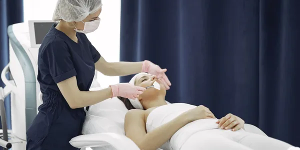 Esteticista de touca e luvas aplicando um tratamento facial em uma cliente deitada na maca