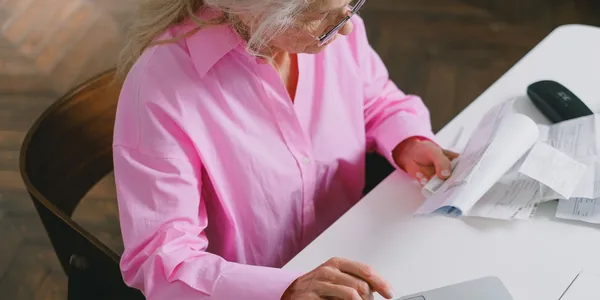 Mulher de camisa rosa conferindo extratos e comprovantes impressos ao lado do notebook aberto sobre a mesa branca