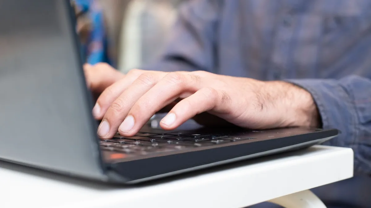 Mãos digitando no teclado de um notebook apoiado em uma mesa branca