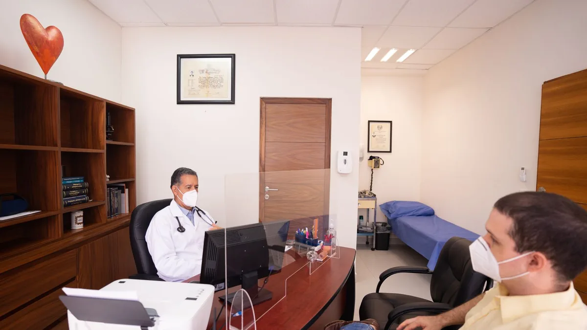 Médica de jaleco branco atendendo uma paciente sentada em um consultório claro