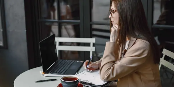 Mulher sentada à mesa de casa conferindo recibos e anotando contas ao lado do notebook, rotina de quem cuida da contabilidade para autônomo