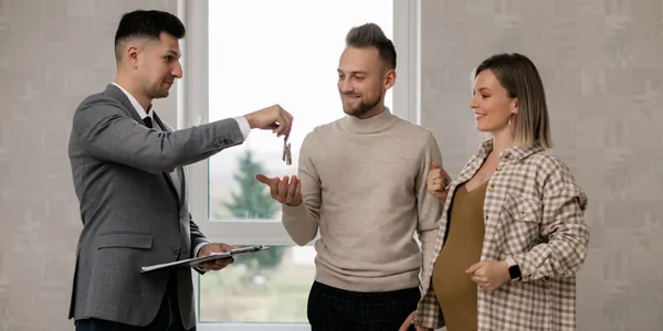 Corretor de imóveis entregando a chave a um casal, com a prancheta do contrato na mão, dentro do apartamento.