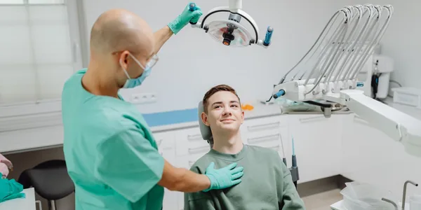 Dentista posicionando o refletor sobre um paciente sentado na cadeira de um consultório odontológico claro.