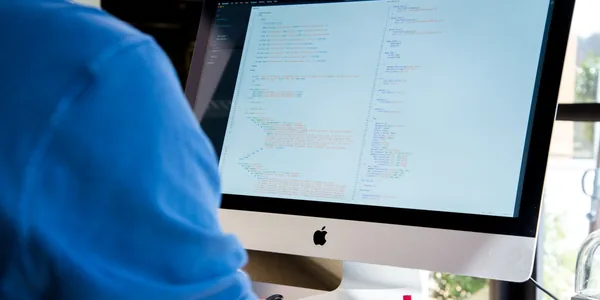 Pessoa de camisa azul programando em um computador de mesa com código na tela.