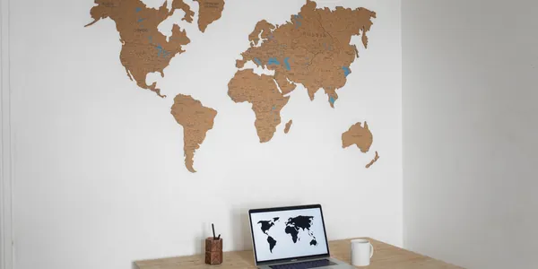 Mesa de trabalho com notebook aberto exibindo um mapa-m&uacute;ndi, diante de um mapa do mundo na parede