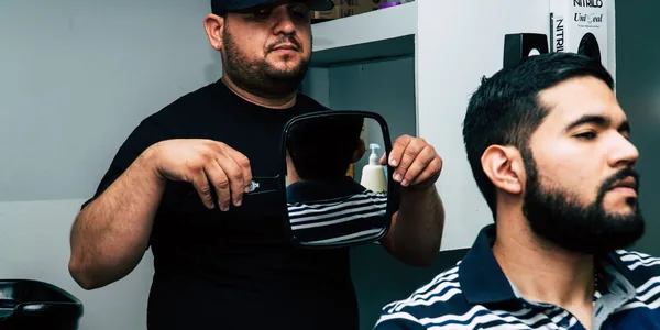 Barbeiro mostrando o corte no espelho para um cliente sentado na cadeira da barbearia.
