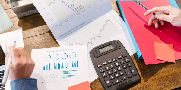 M&atilde;o apontando para gr&aacute;ficos financeiros impressos sobre a mesa, ao lado de uma calculadora.