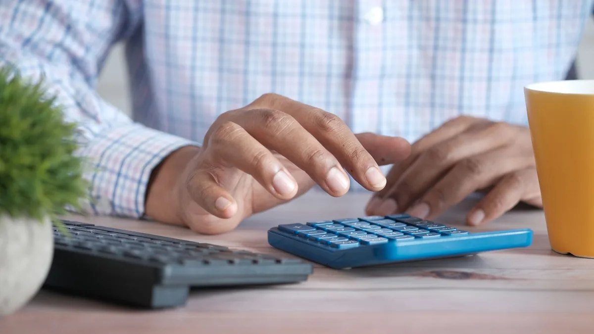 Mãos digitando números em uma calculadora sobre a mesa de trabalho, ao lado do teclado do computador.