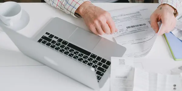 Pessoa conferindo notas fiscais impressas com código de barras e QR Code ao lado do notebook em uma mesa de escritório