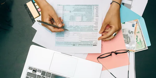 Mãos preenchendo documentos de imposto ao lado de um notebook e óculos